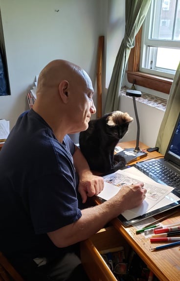Jay sits at his desk colouring a small saves cartoon as his cat, Sedona, watches